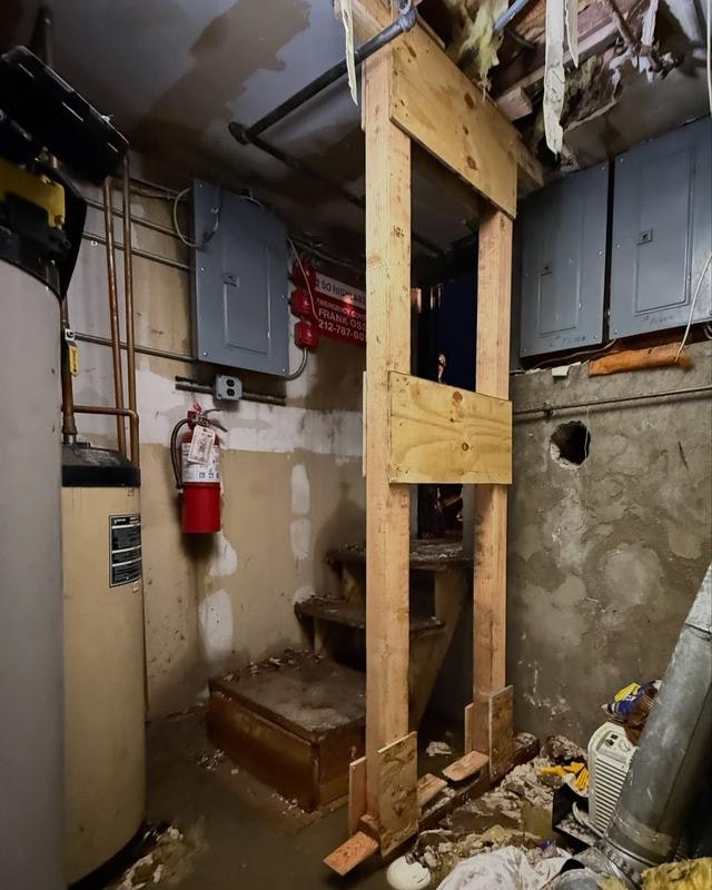 The image depicts a basement area with wooden supports, stairs, electrical panels, and some clutter, including a water heater and fire extinguisher.