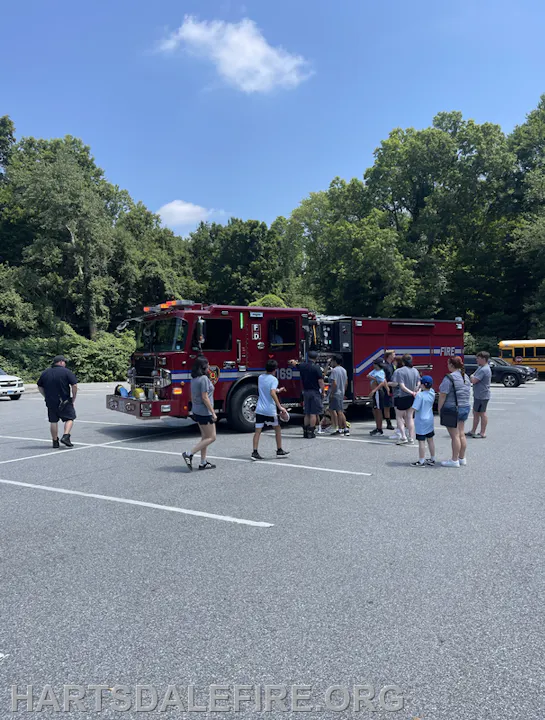 A group of kids gathers around a fire truck on a sunny day in a parking lot, with trees in the background.