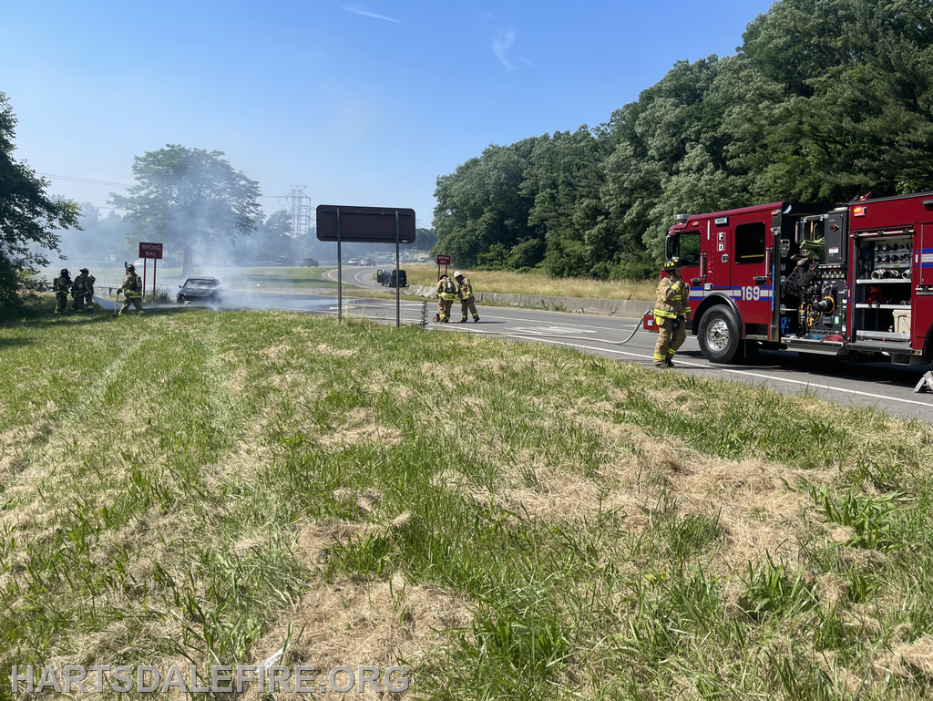Firefighters are responding to a car fire on the side of a road, with smoke visible and a fire truck present.