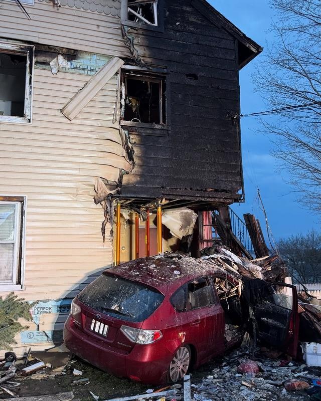 The image shows a fire-damaged house, with severe charring on one side and a red car wrecked against its base.