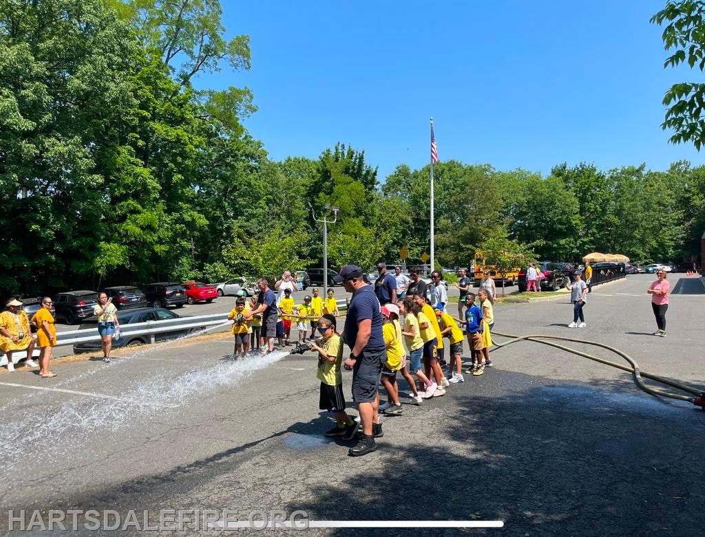 A group of children in yellow shirts spray water with a hose outside, supervised by adults on a sunny day.