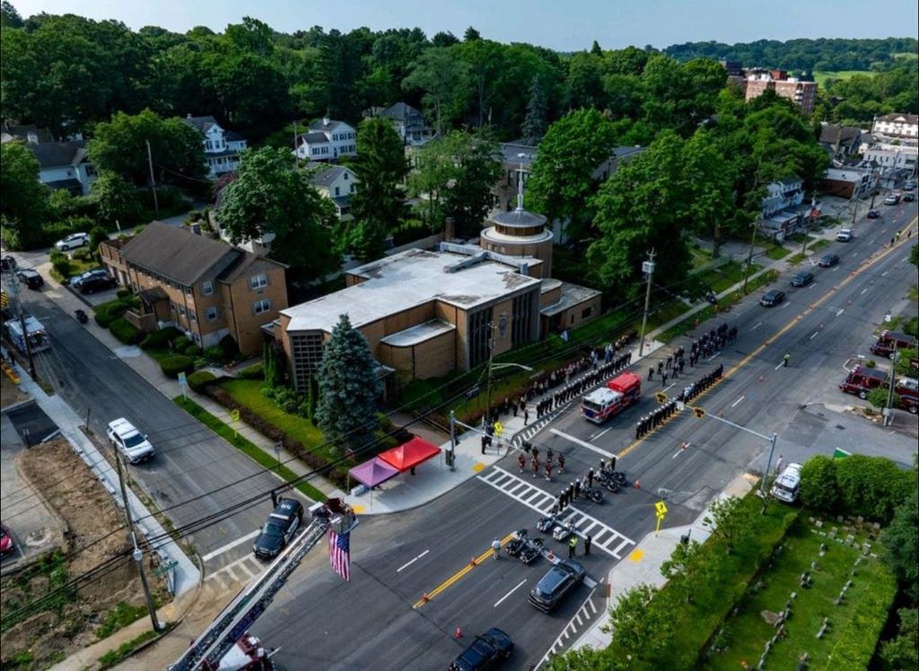 An aerial view of a procession with vehicles, people lined up, and a building, set in a suburban area surrounded by trees.