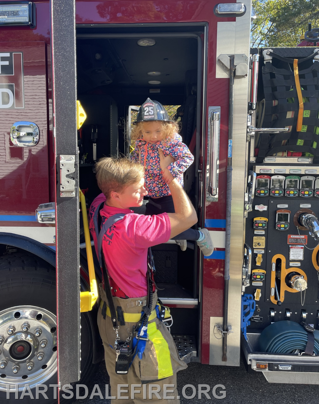 A firefighter helps a young girl wearing a toy fireman's hat get out of a fire truck.