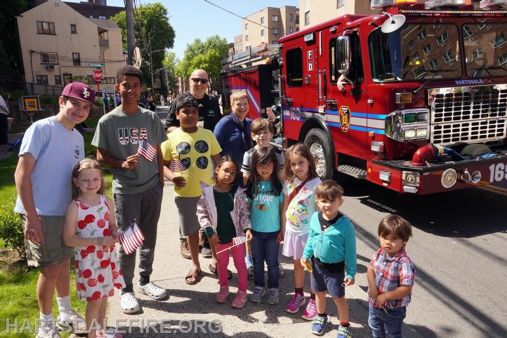 A group of children and adults gather with small American flags in front of a firetruck on a sunny day.