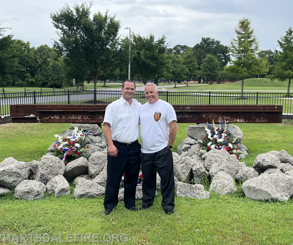 Two men stand together near a memorial with flowers and rocks, featuring a steel beam in the background. It looks like a tribute site.