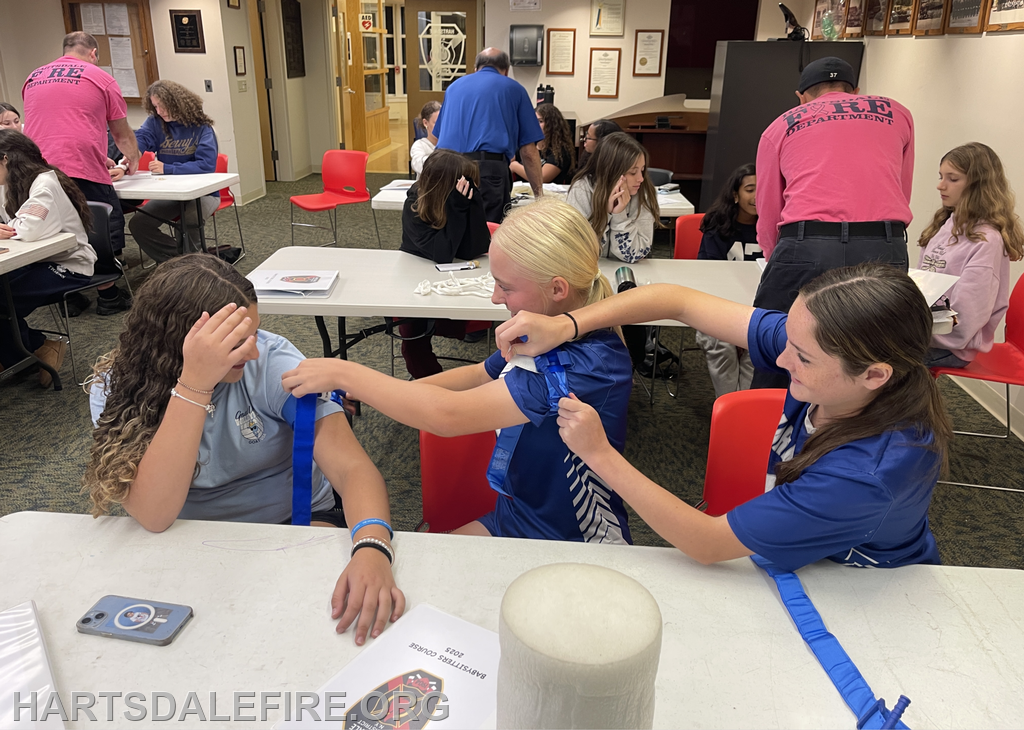 A group of young people practicing emergency techniques, including bandaging, in a classroom setting.
