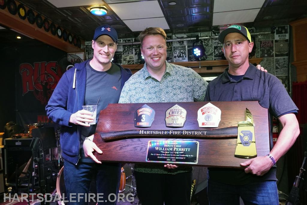 Three men pose with a wooden plaque honoring Deputy Chief William Perritt from Hartsdale Fire District at an event.