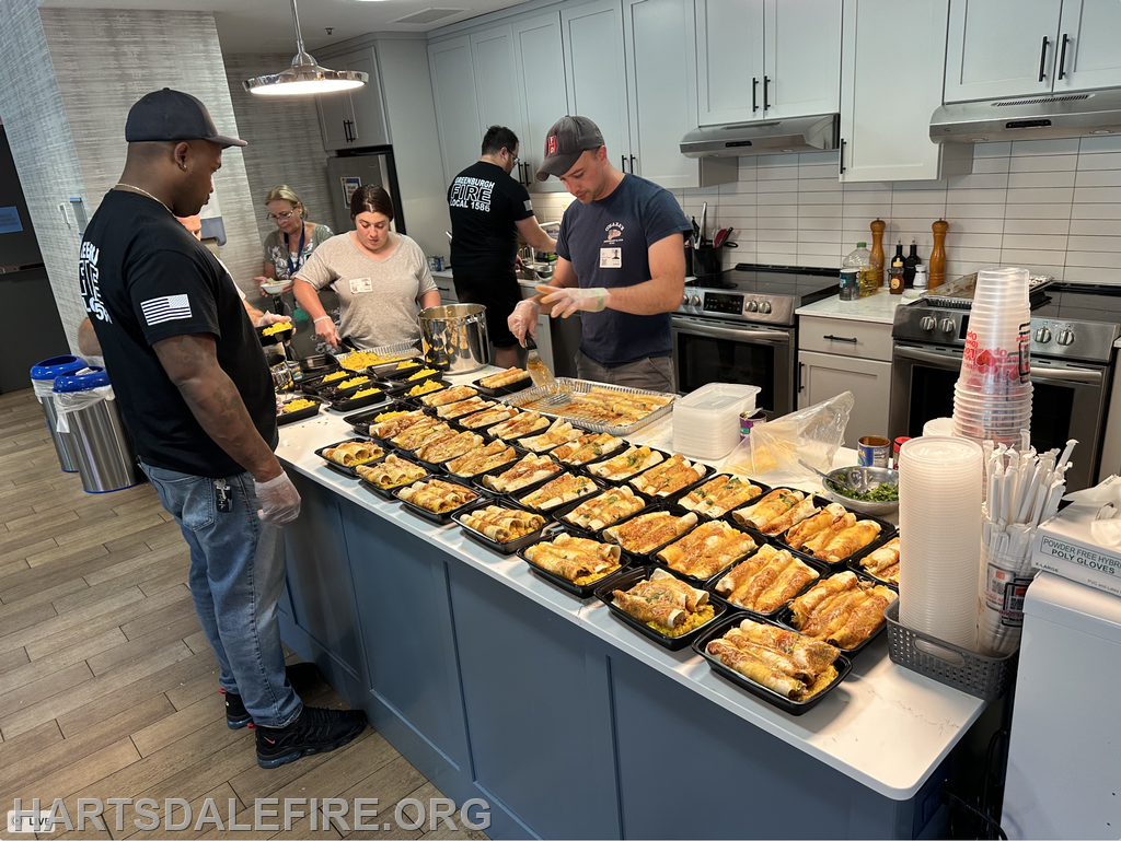 A group is preparing food in a kitchen, with several trays of meals arranged on a counter.
