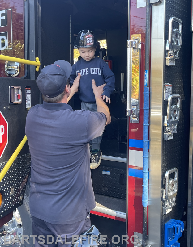 A firefighter assists a young child, wearing a helmet, as he steps off a fire truck.
