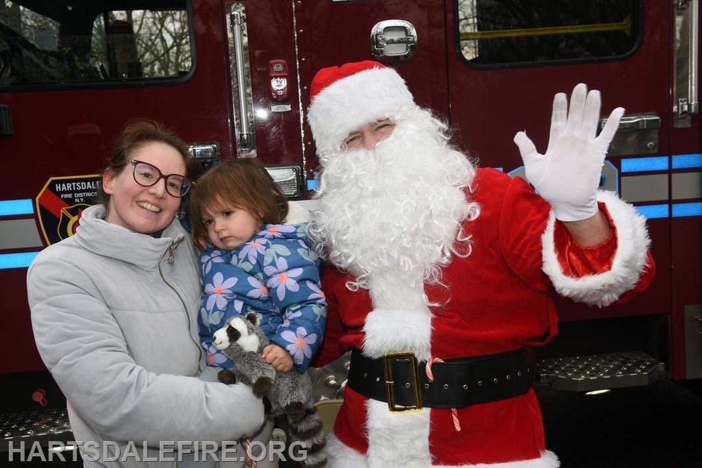 A woman and a young girl pose with Santa Claus in front of a fire truck, capturing a festive moment.