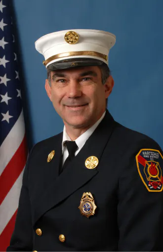 The image features a man in a formal fire service uniform, wearing a white hat, posed in front of an American flag backdrop.