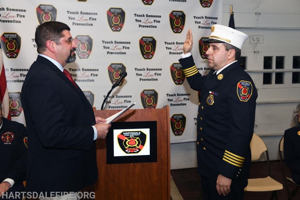 A fire official is being sworn in, raising hand, next to a podium with a backdrop of fire department logos.