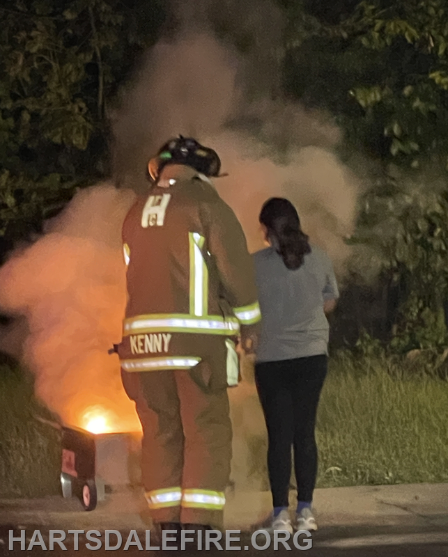 A firefighter in gear stands next to a woman, both observing smoke and flames coming from a cart.