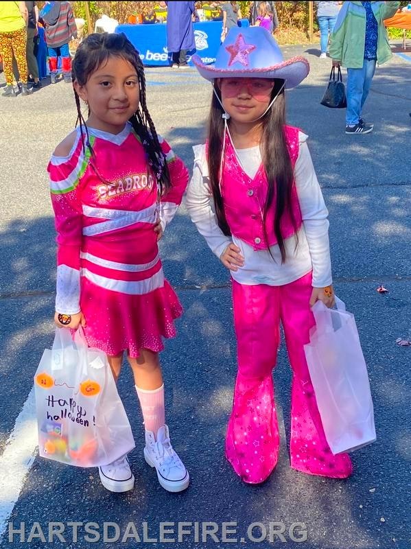 Two girls in colorful Halloween costumes pose for a photo, each holding a bag of treats.