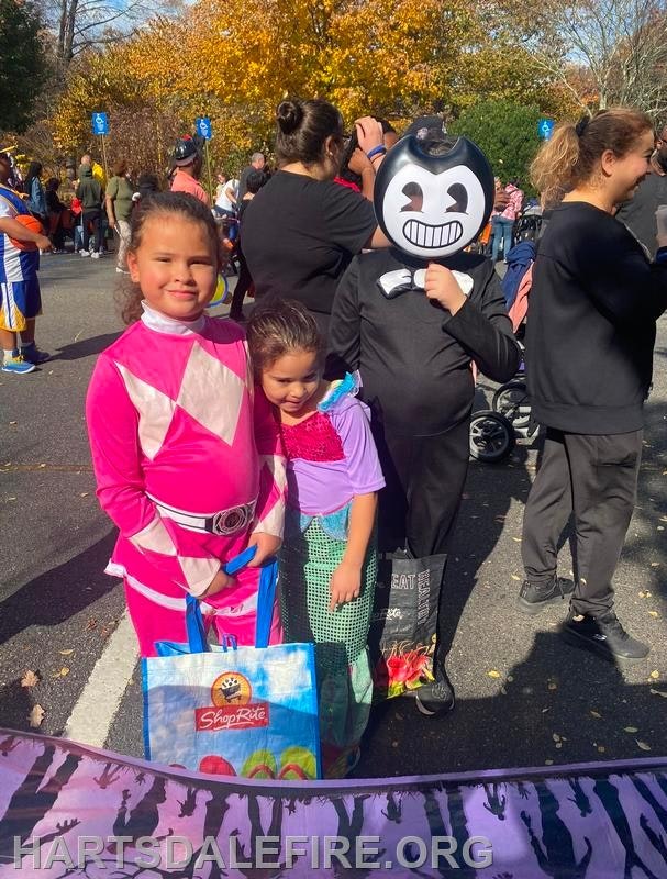 Three children in costumes pose for a photo during a festive outdoor event, with colorful bags and autumn foliage in the background.