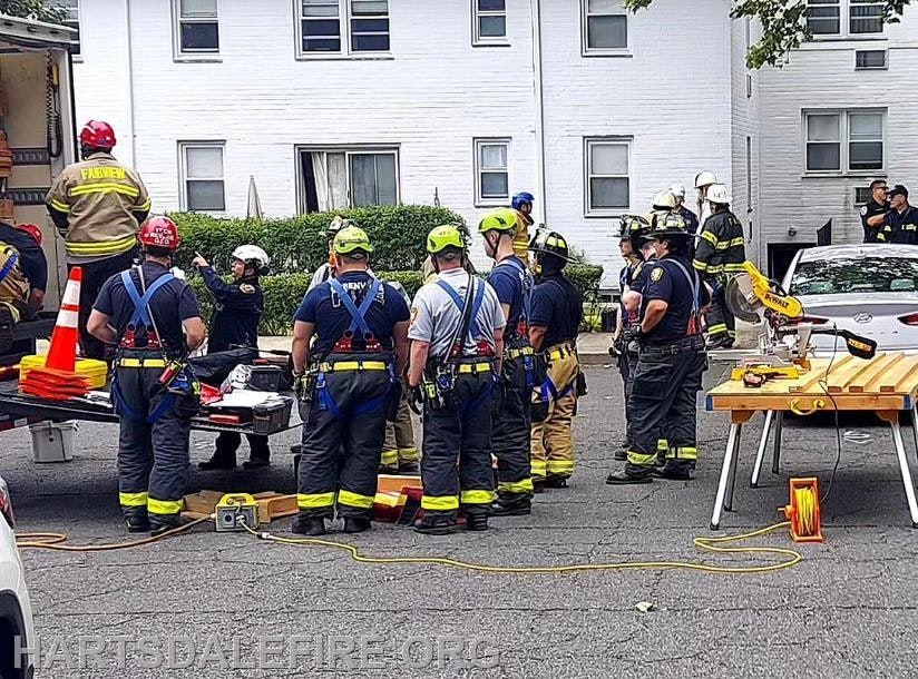 Firefighters in gear stand near a truck with equipment, in front of a white building during a training or emergency response event.