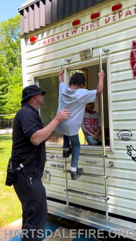 A firefighter assists a child climbing a ladder into a fire department trailer.