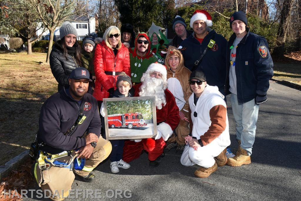 A group of people, including Santa, gather outdoors for a festive photo, holding a picture of a fire truck.