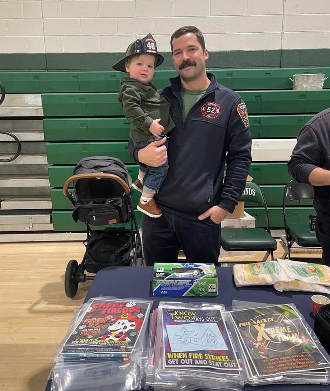 A fireman holds a toddler wearing a mini fire hat at an event, surrounded by fire safety pamphlets and a stroller.