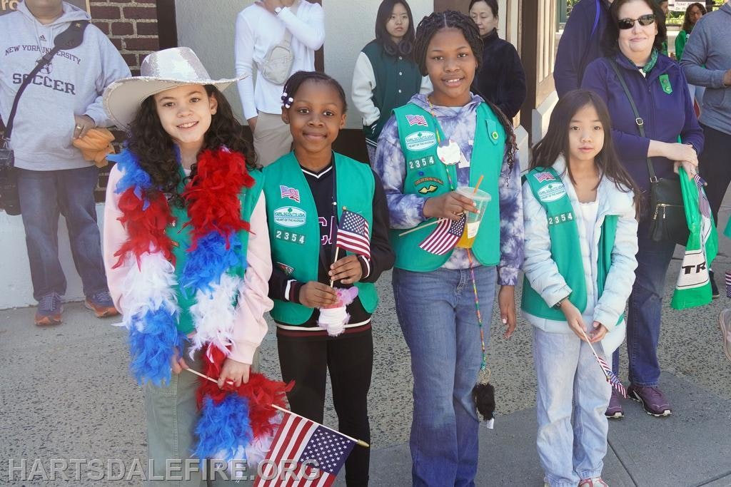The image shows a group of girls in green vests, holding American flags and celebrating, likely at a parade or event.