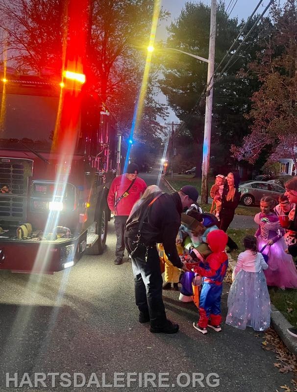 A fire truck is parked as children in costumes interact with firefighters during a festive evening event.