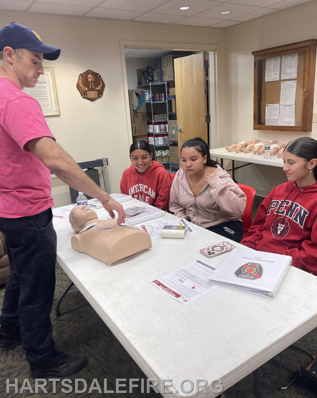 A trainer demonstrates on a CPR dummy while three young learners watch attentively, engaged in a learning session.