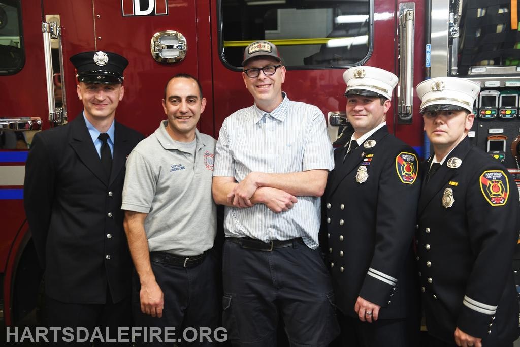 Five people, including firefighters, pose in front of a fire truck; one wears casual clothing, the others in uniforms.