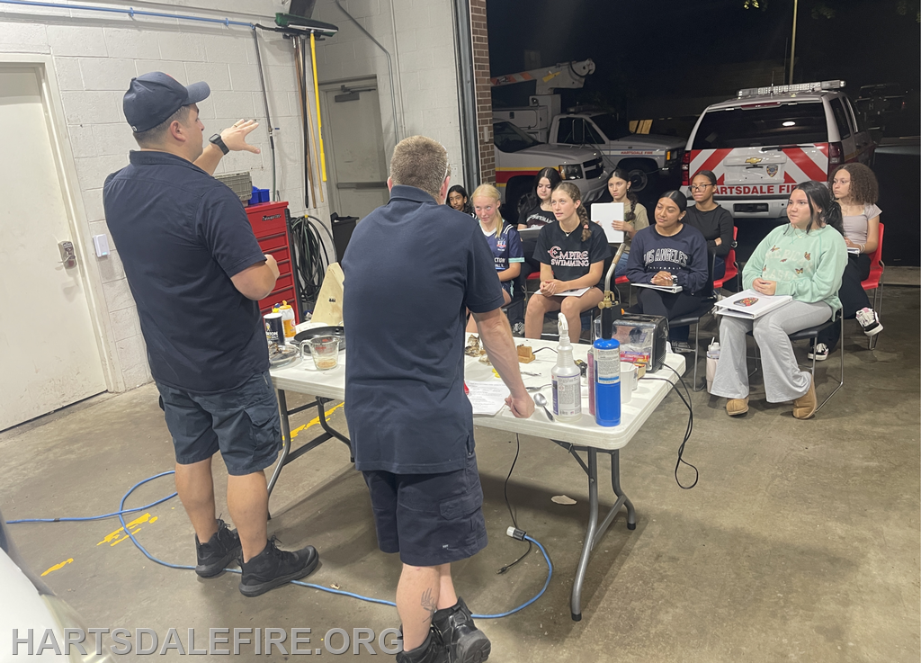 Two instructors teach a group of students in a firehouse, discussing safety procedures with tools and materials on a table.