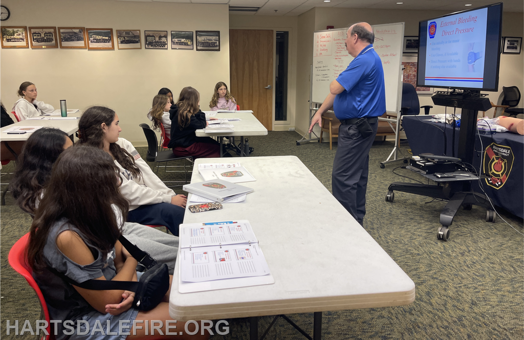 A group of young people listens to a presentation on emergency care, with materials and a demonstration setup visible.
