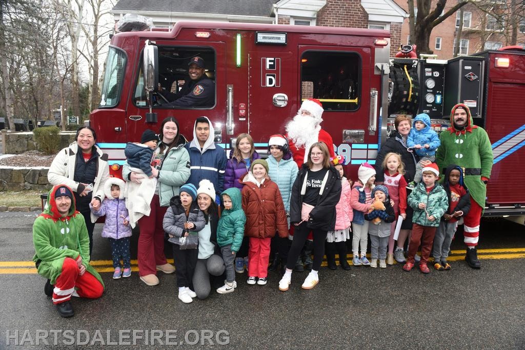 A festive group poses with Santa, elves, and a fire truck during a community holiday event.
