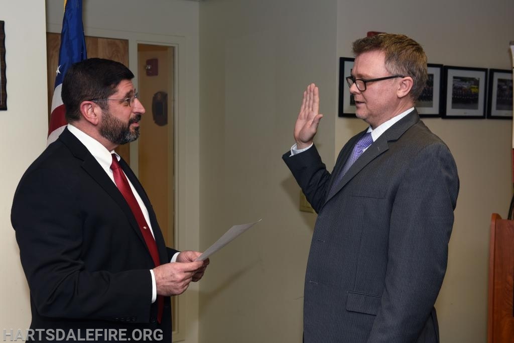 Two men in formal attire, one holding a paper, the other raising his right hand, in an indoor setting.