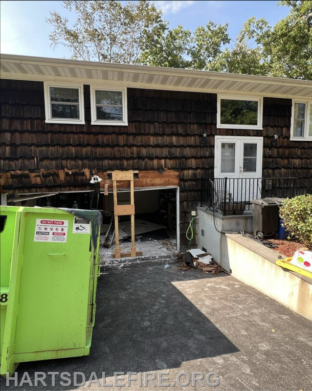 A house with wooden shingle siding, a construction area, and a green dumpster in front, suggesting ongoing renovation work.