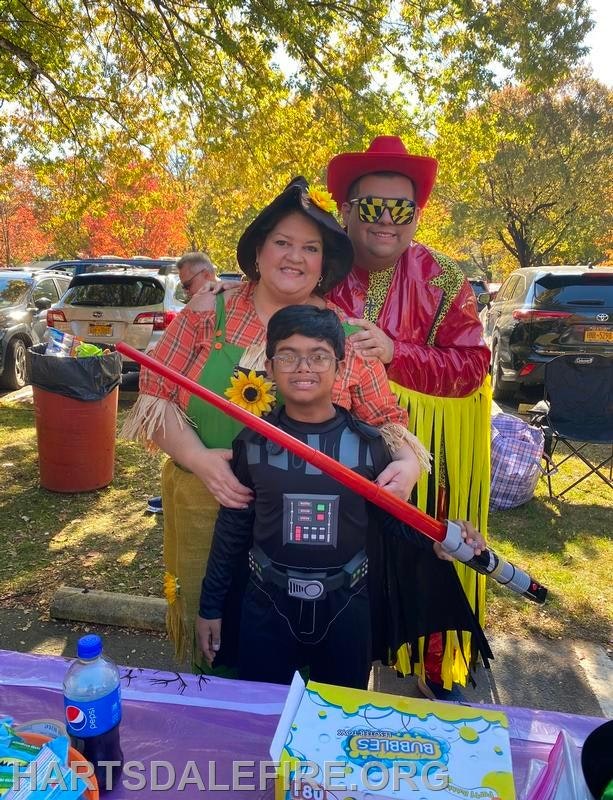 A group in costumes poses together outdoors: a child dressed as Darth Vader holds a lightsaber, flanked by two adults in colorful attire.