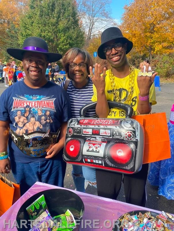 Three people are smiling at a lively outdoor event, with one holding a large boombox and wearing fun accessories.