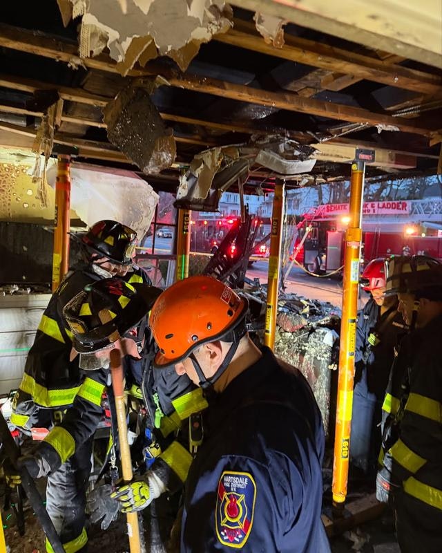 Firefighters work under damaged ceiling structures, responding to an emergency situation with fire trucks visible outside.