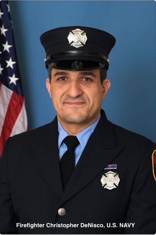A uniformed firefighter poses for a portrait against a blue background, with an American flag visible behind him.