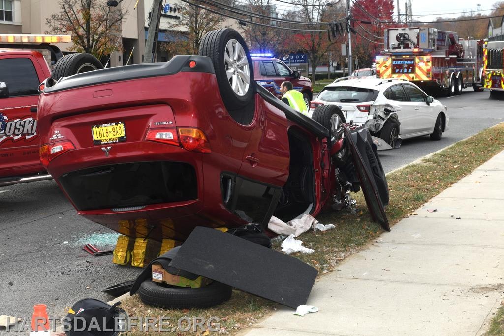 A red car is overturned on its side near a crash scene, with emergency vehicles and a damaged white car visible nearby.