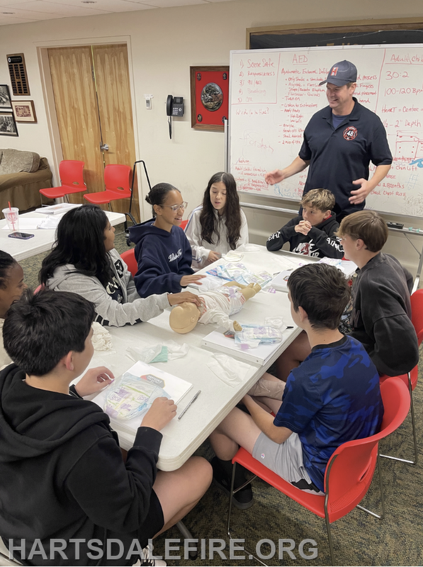 A group of children learning CPR with an instructor, using a dummy in a classroom setting.