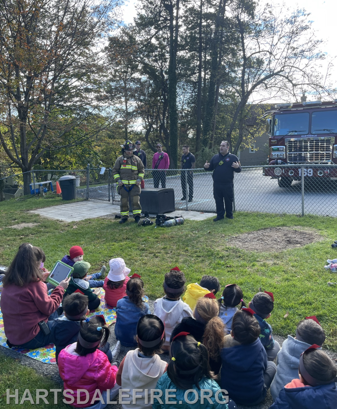 A firefighter and a speaker educate a group of children about fire safety near a fire truck, promoting community awareness.