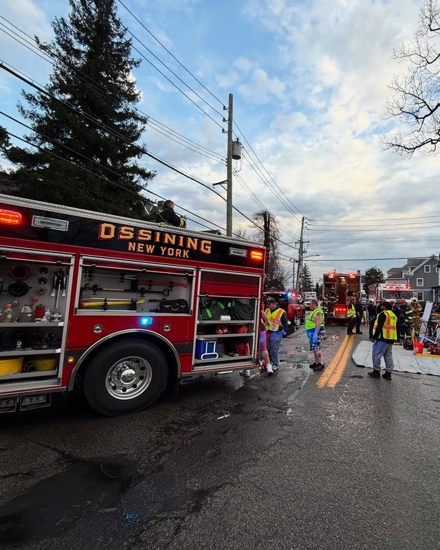 Emergency response scene with a fire truck labeled "Ossining, New York" and first responders on a street, cloudy sky above.