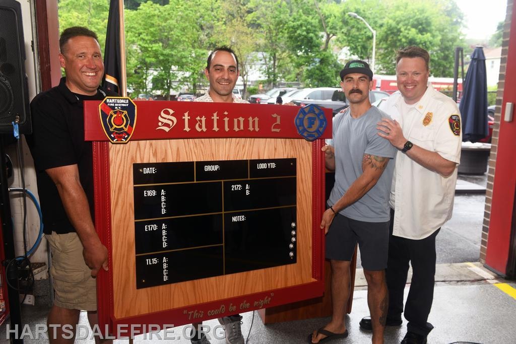 Four individuals are celebrating with a new display board at a fire station, showing a friendly community event.