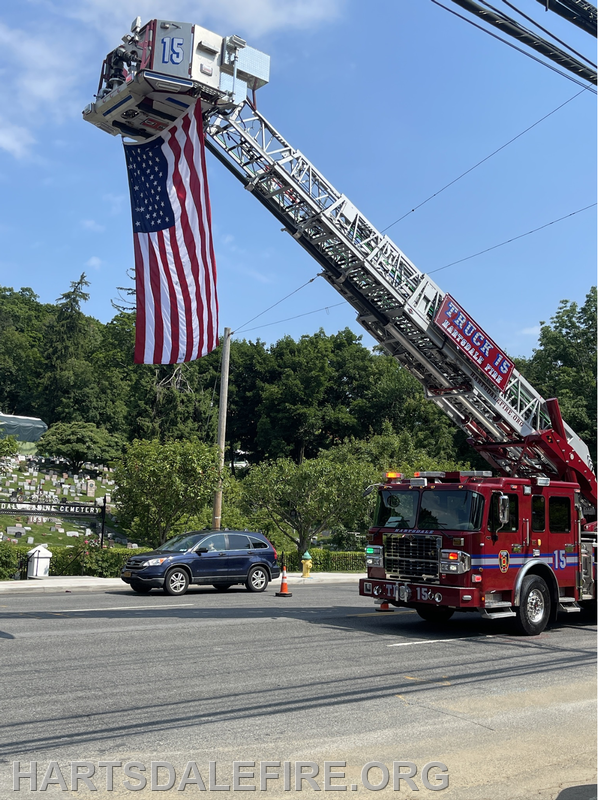 A fire truck extends a ladder displaying the American flag against a blue sky. Cars pass by in a peaceful scene.