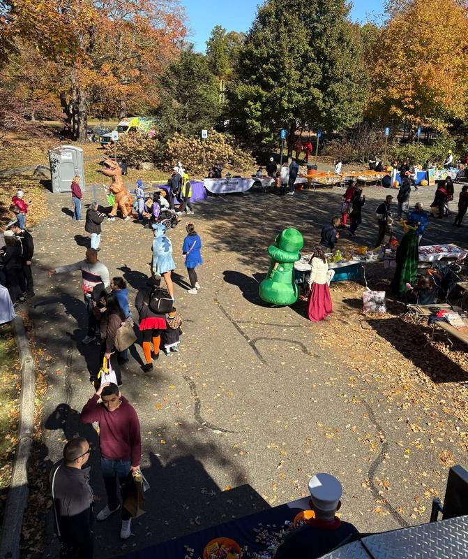 A lively outdoor event with people in costumes, booths, and autumn foliage, creating a festive atmosphere.