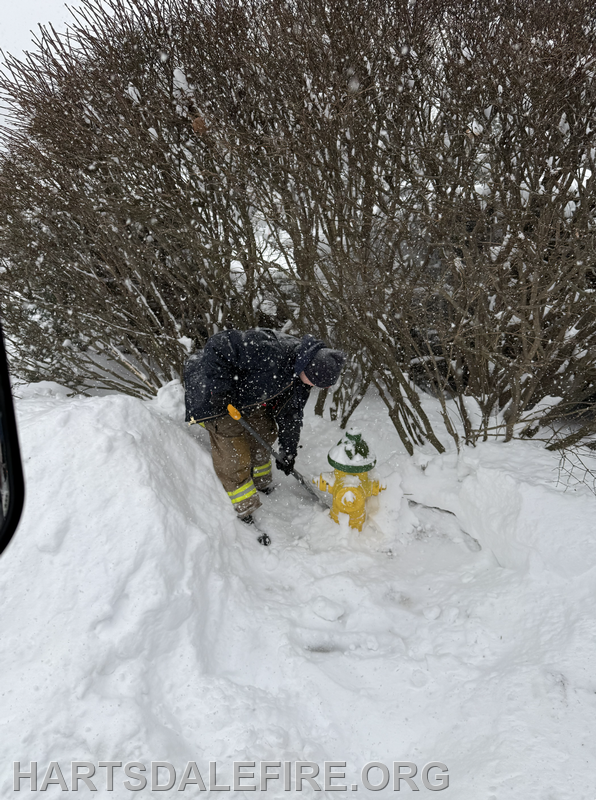 A firefighter is shoveling snow around a yellow fire hydrant during a snowstorm.