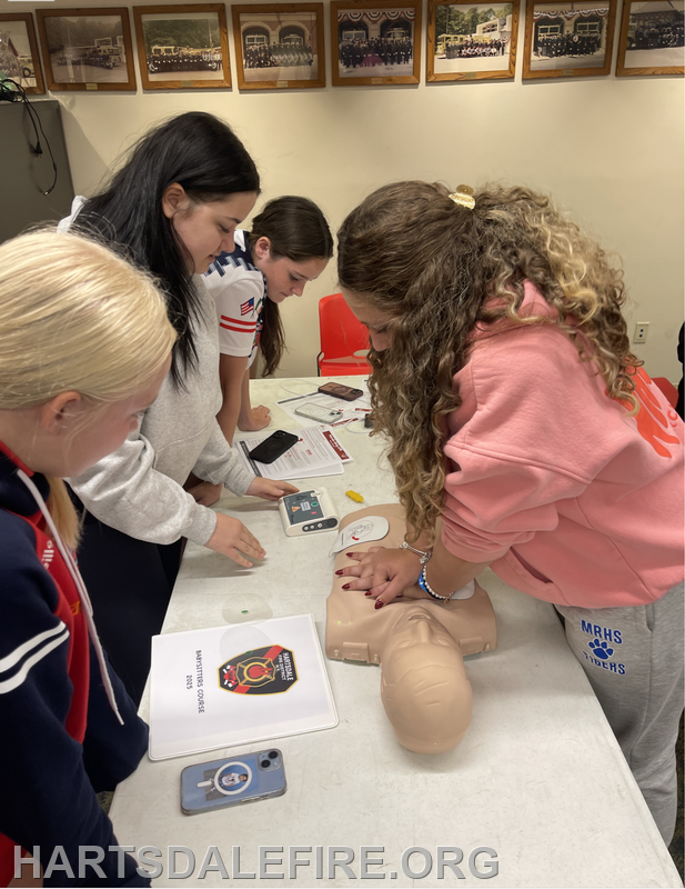 Group of young women practicing CPR techniques on a training dummy, with instructional materials and an AED device on the table.