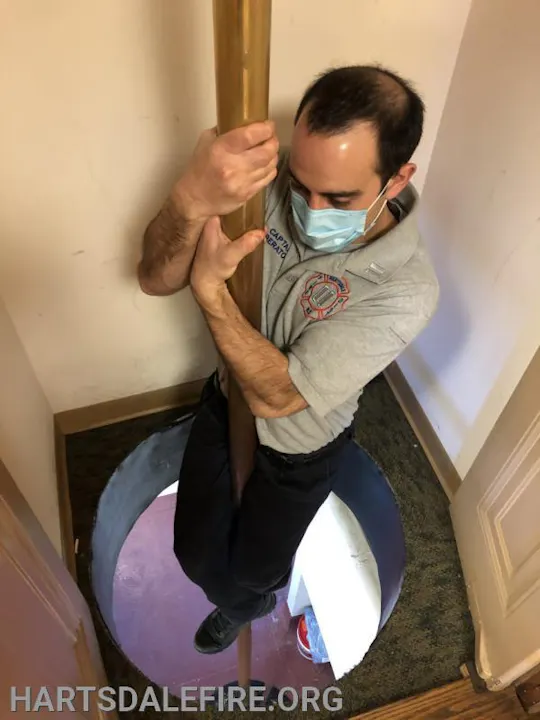 A person wearing a mask slides down a fire pole in a confined indoor space.