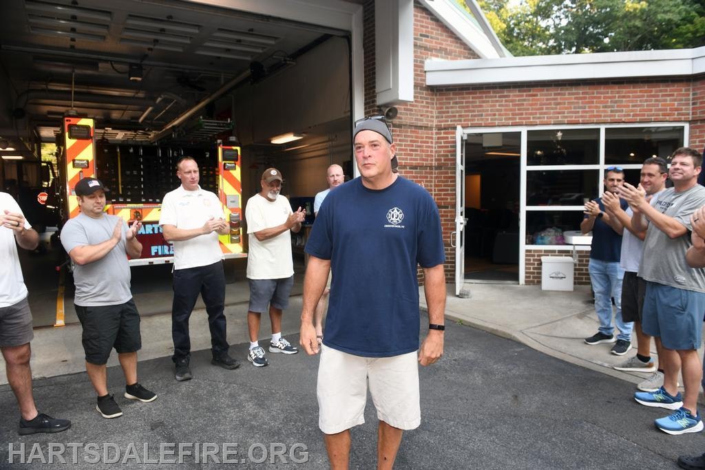 A group of people is applauding a man in a fire station, celebrating him in front of a fire truck.