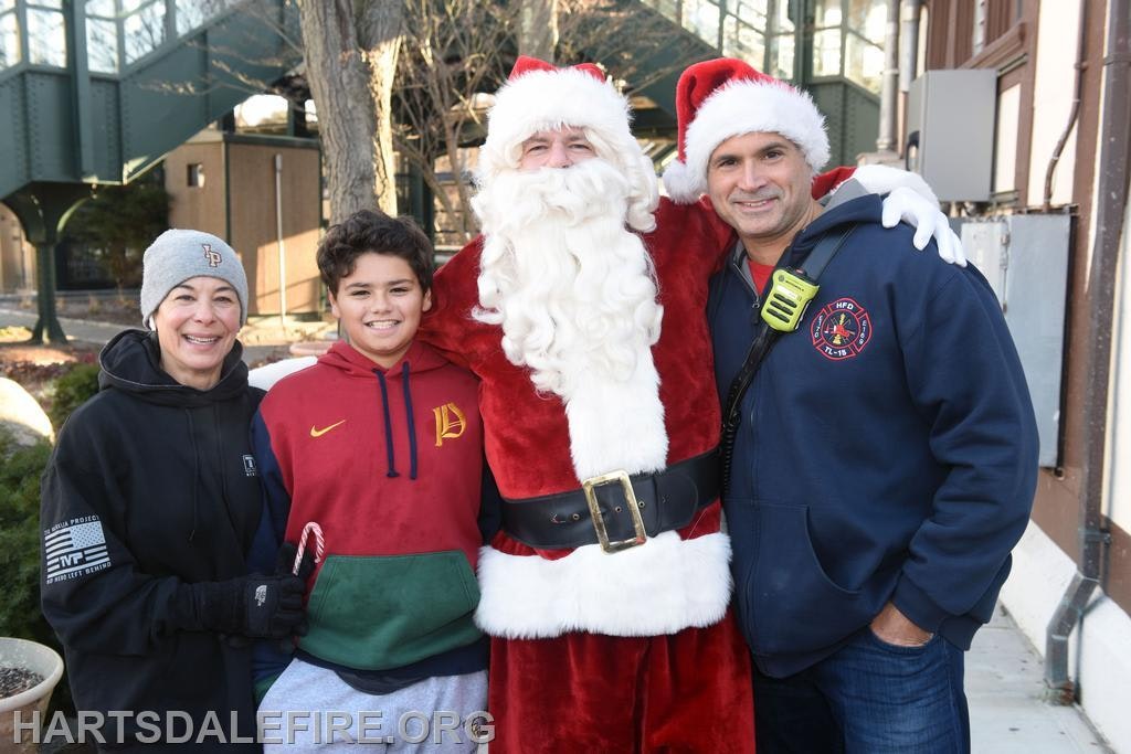 A festive group photo featuring Santa Claus with a family and a firefighter outside, all smiling together.