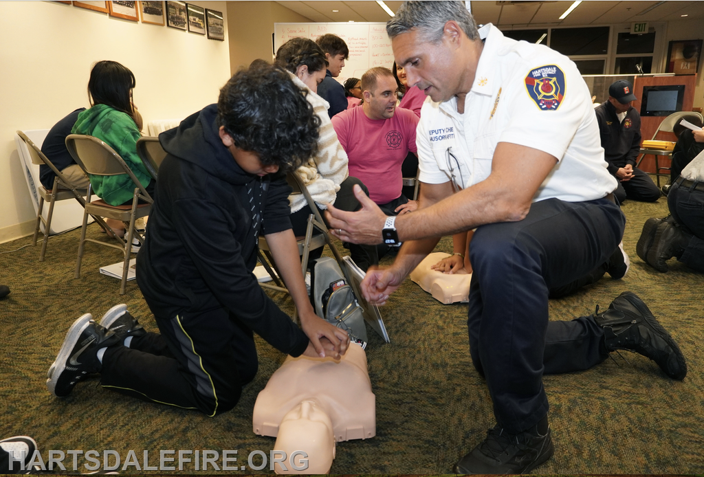 A youth practices CPR on a training dummy, guided by a firefighter, in a classroom setting. Other participants observe.