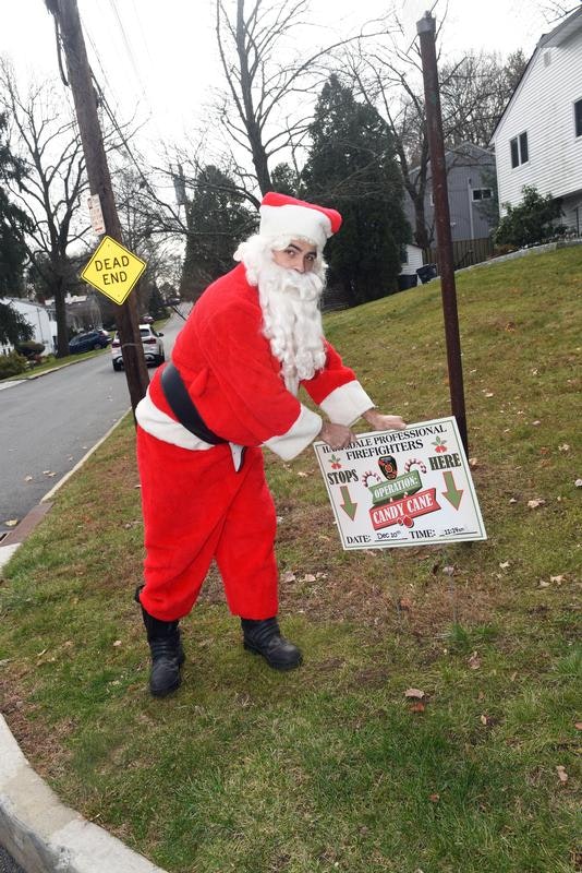 Santa holding a sign for a firefighter event called "Operation Candy Cane" on the grass near a "Dead End" road sign.